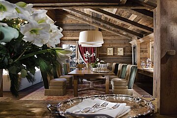 A cozy, rustic wooden dining room in a chalet, featuring a long table, upholstered chairs, white flowers, and a silver tray with cutlery in the foreground.