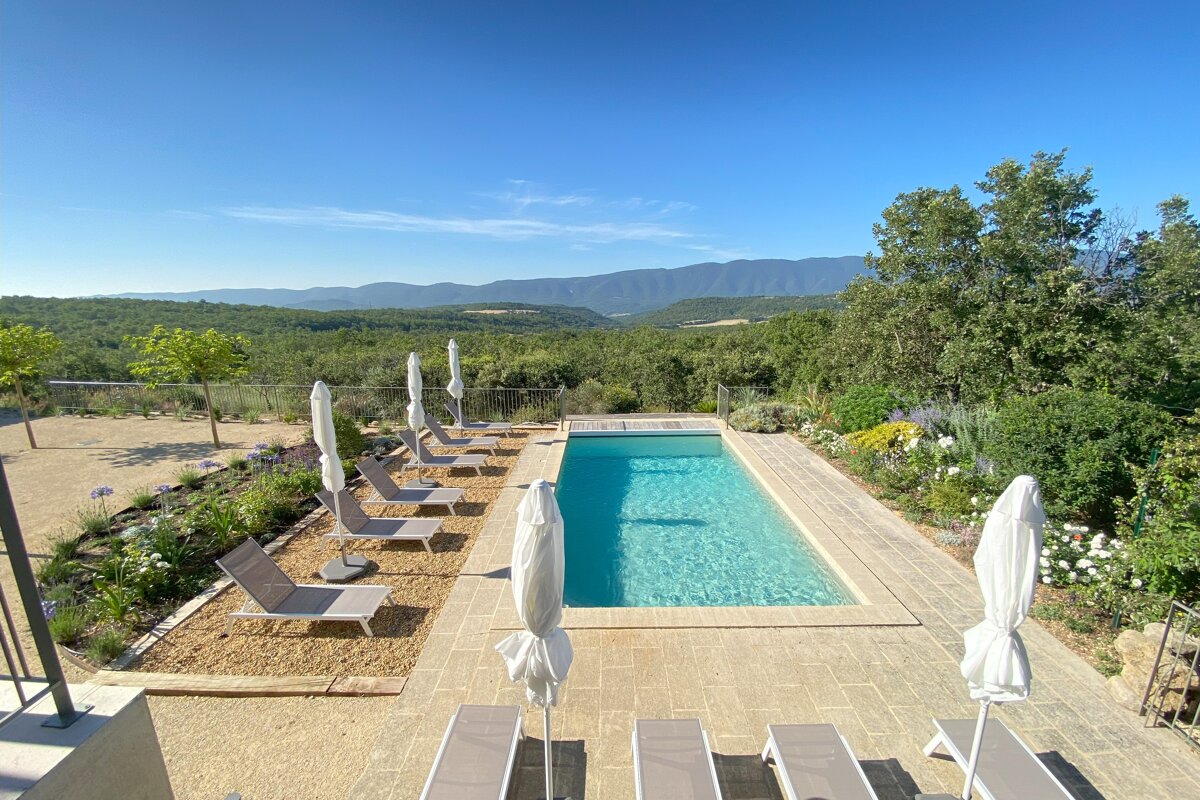A serene outdoor swimming pool with lounge chairs and umbrellas, overlooking a vast landscape of lush green forests and mountains under a clear blue sky.
