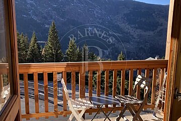 A wooden balcony with chairs & table offers a scenic view of pine trees, a road, and majestic mountains under a clear sky.