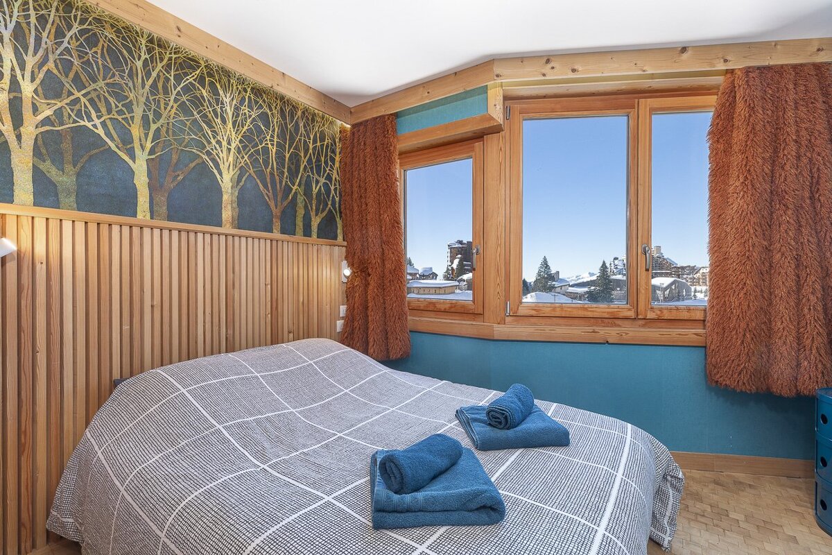 A cozy mountain bedroom with a checkered bed, tree-patterned wallpaper, and snowy views through wooden windows.