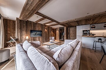 Cozy, modern alpine chalet interior with rustic wood-paneled walls, a large grey sectional sofa, and an open kitchen area.
