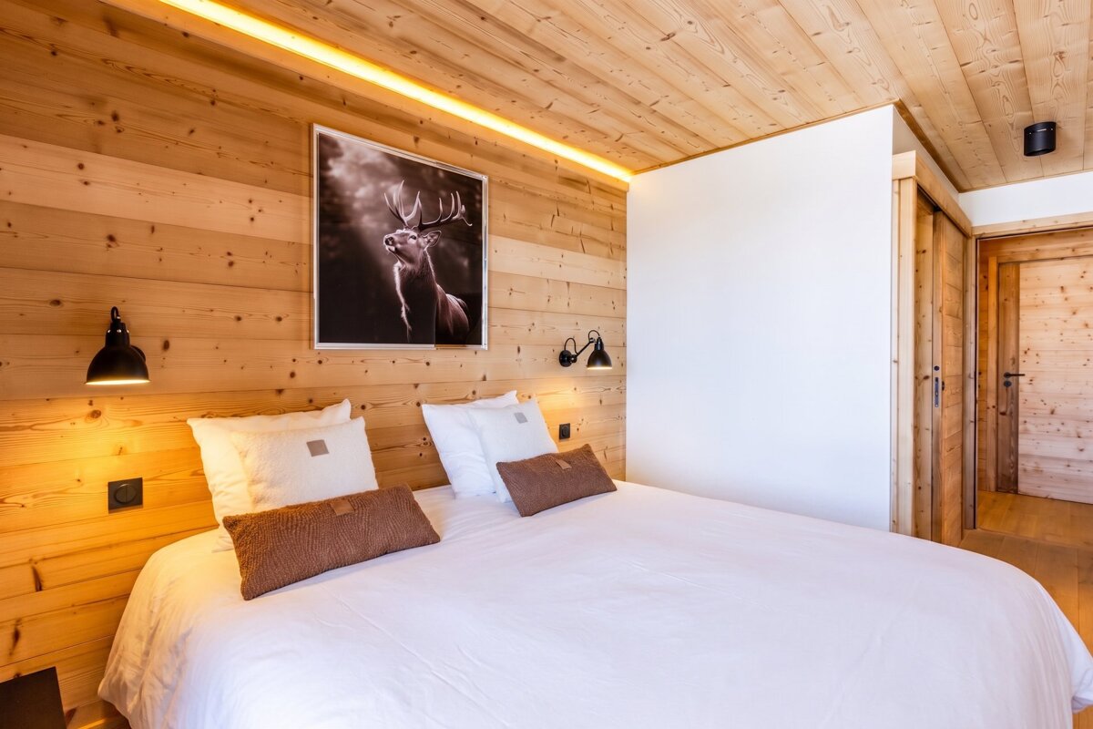 Cozy wooden chalet bedroom featuring a bed with white linen, black bedside lamps, and a framed picture of a stag on the paneled wall.