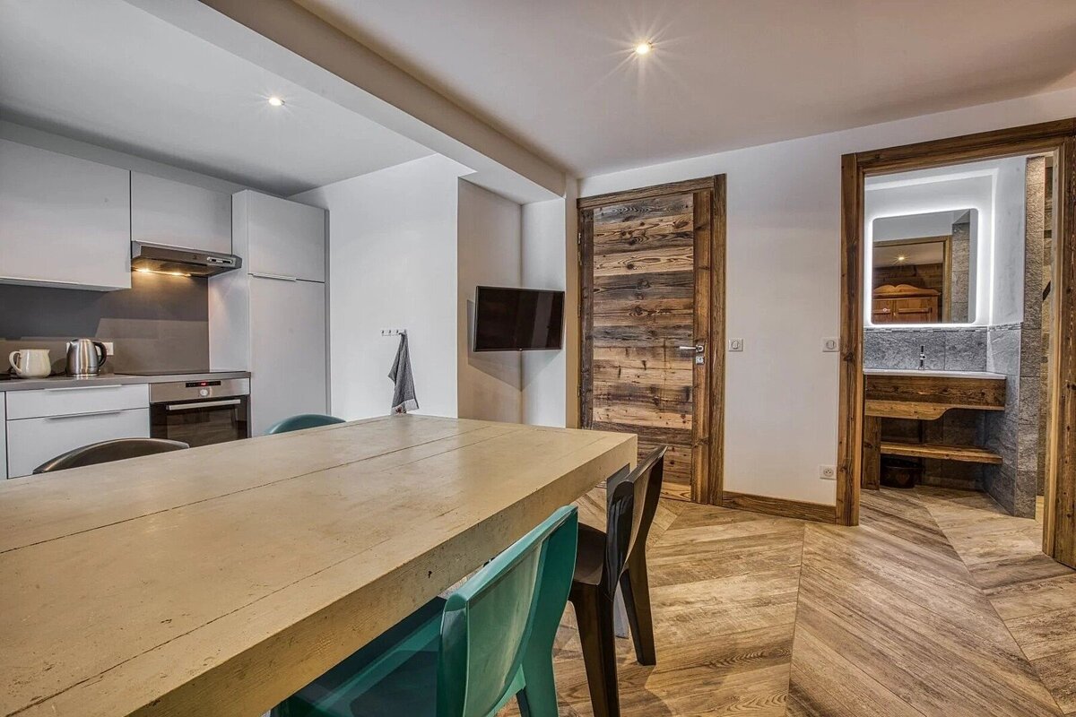 A modern kitchen/dining area with a wooden table, contrasted by rustic wooden doors and bathroom entrance. Warm wood-patterned floors complete the look.