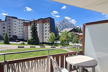 Balcony view of a mountain resort with modern buildings, a grassy lawn, and a scenic peak under a bright blue sky.