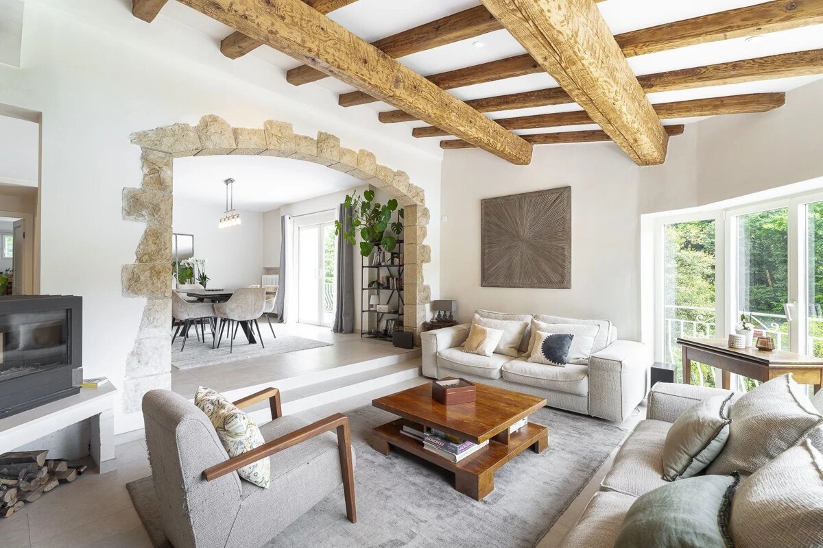 Modern rustic living room featuring exposed wooden ceiling beams, a stone archway, and neutral sofas with a wooden coffee table.