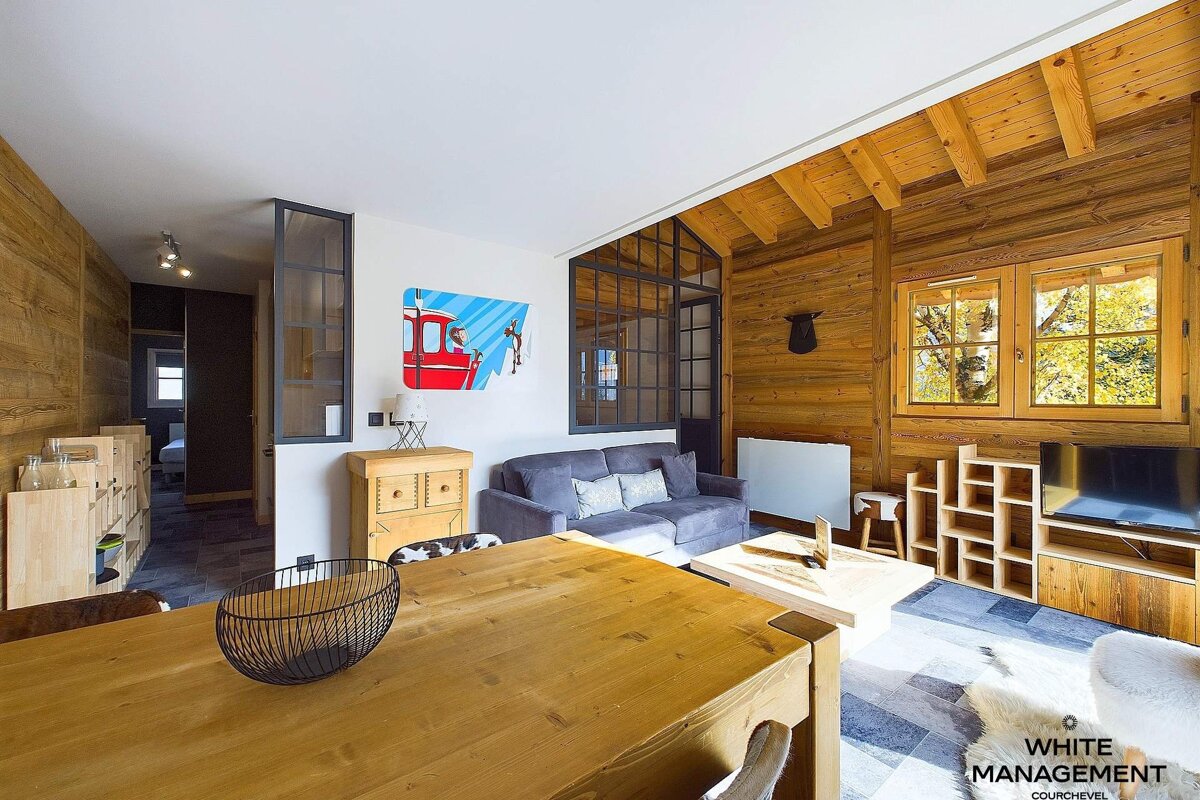 A cozy, rustic-modern ski chalet living and dining room with extensive wood paneling, a large wooden table, gray sofa, and colorful gondola art.