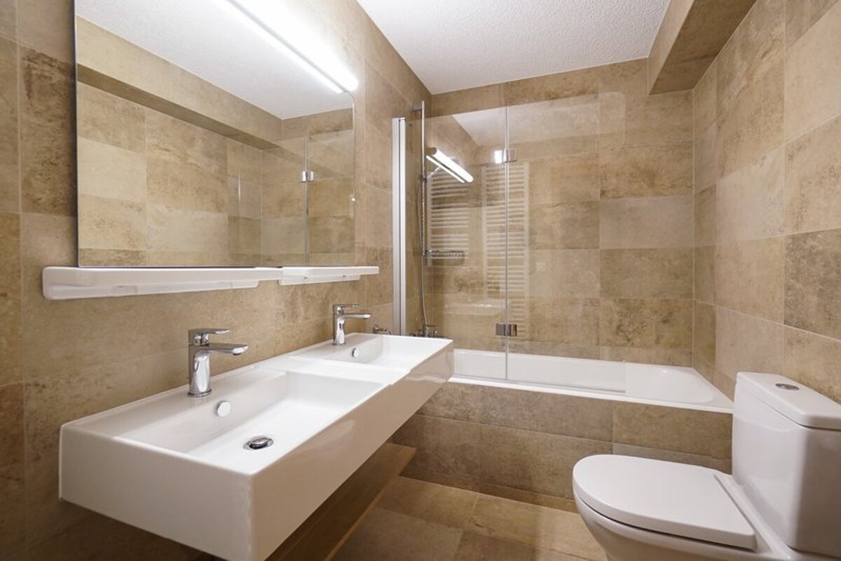 Modern bathroom with beige tiling, featuring a double sink vanity, a toilet, and a bathtub with a glass screen.