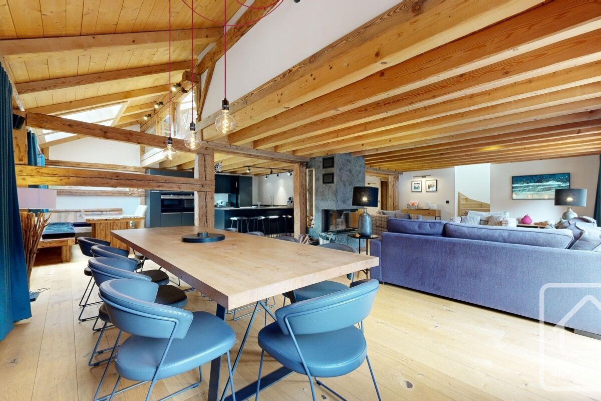 Modern, spacious open-plan room with exposed wooden beams, light wood floors, a dining area, cozy lounge with a fireplace, and a contemporary kitchen.