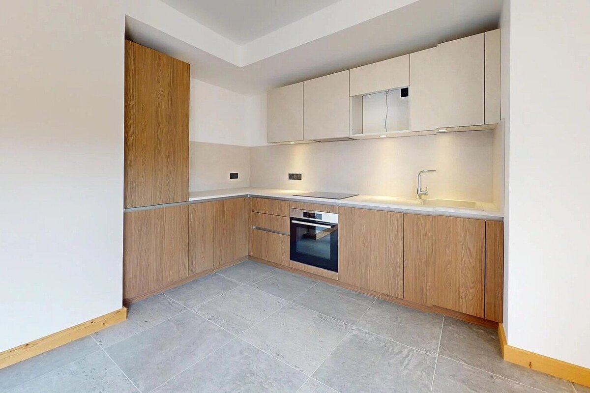 A modern L-shaped kitchen featuring wood grain base cabinets, light upper cabinets, integrated oven, under-cabinet lighting, and gray tiled floor.