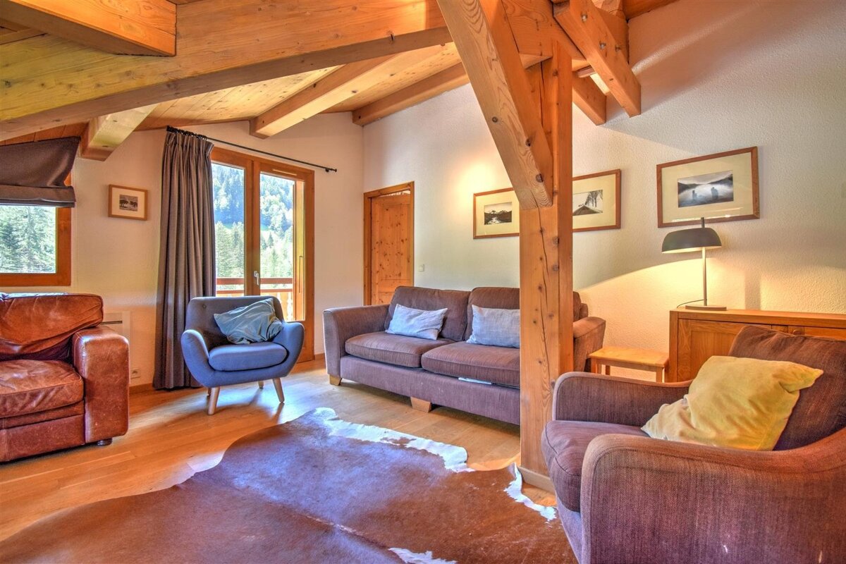 A cozy, rustic living room with exposed wooden beams, a cowhide rug, and various seating, overlooking a forested mountain view from a balcony.