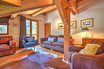 A cozy chalet living room with exposed wooden beams, various seating, a cowhide rug, and a balcony door revealing a mountain view.