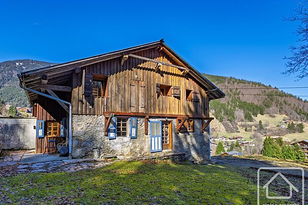 Rustic wooden and stone house with blue shutters on a sunny day, set against a backdrop of green mountains.