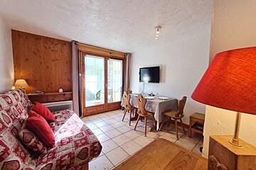 A small living/dining room featuring a red patterned sofa bed, wood-paneled wall, dining table with four chairs, wall TV, and glass patio doors.