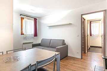 A bright, modern living/dining area featuring a grey sofa, dining table, light wood floor, white walls, and a window with red and white curtains.