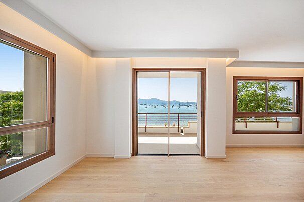 An empty, modern room with light wooden floors and large windows, one with a sliding glass door opening to a balcony with a sea view and boats.