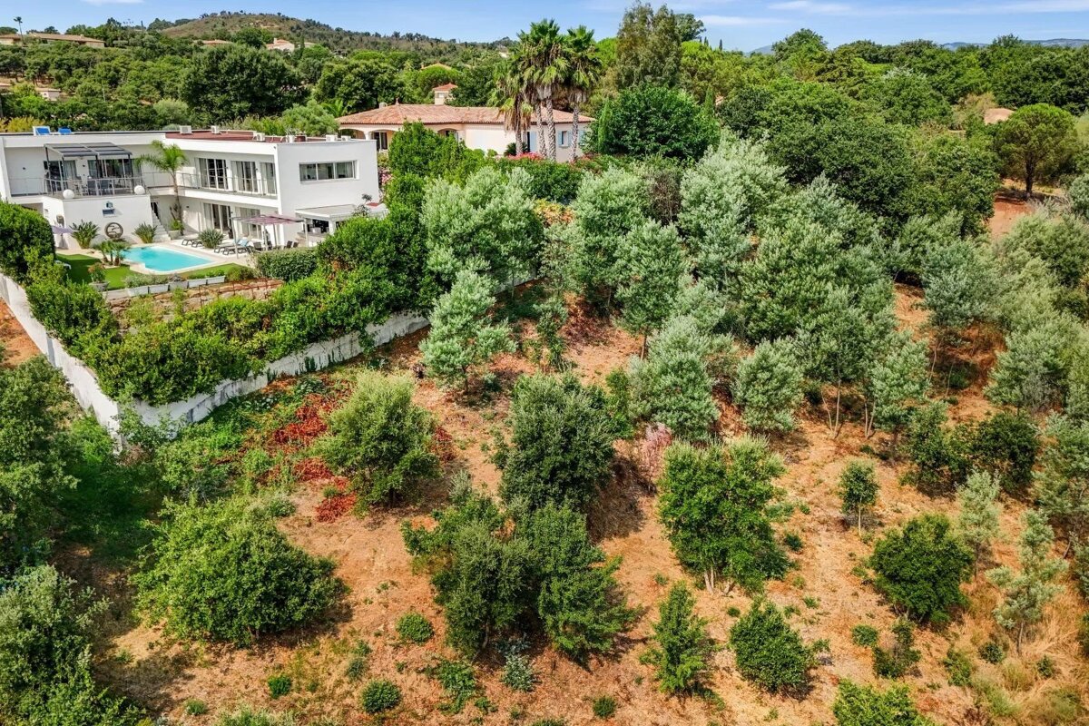 Aerial view of a modern white villa with a pool, surrounded by lush green trees and a hillside on a sunny day.