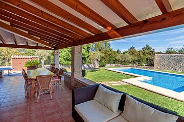 A covered patio with dining and lounge areas overlooks a sunny green lawn and a sparkling blue swimming pool, surrounded by lush trees.