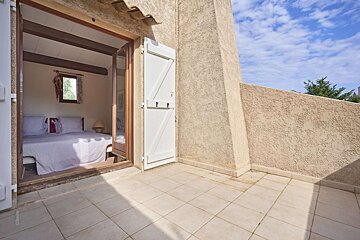 A bedroom with a gingham bedspread seen through open French doors leading to a tiled balcony under a blue sky, flanked by textured walls.