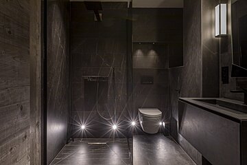 A minimalist, dark marble bathroom featuring a walk-in shower, wall-hung toilet, sleek vanity, and dramatic low-level spotlighting.