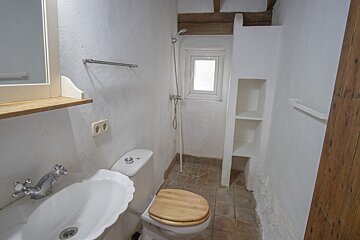 A rustic white bathroom with a sink, toilet with a wooden seat, a shower, small window, tiled floor, and built-in shelves.