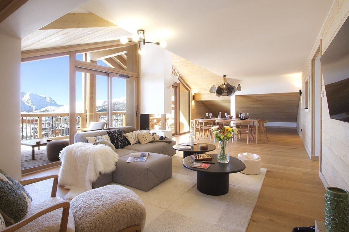 A stylish, open-plan chalet living and dining area with a cozy sofa, elegant decor, and large windows overlooking stunning snowy mountains.