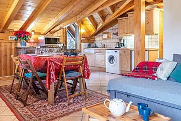 Cozy alpine cabin interior featuring exposed wooden beams, a dining table with a red tablecloth, a small kitchen, and a living area.