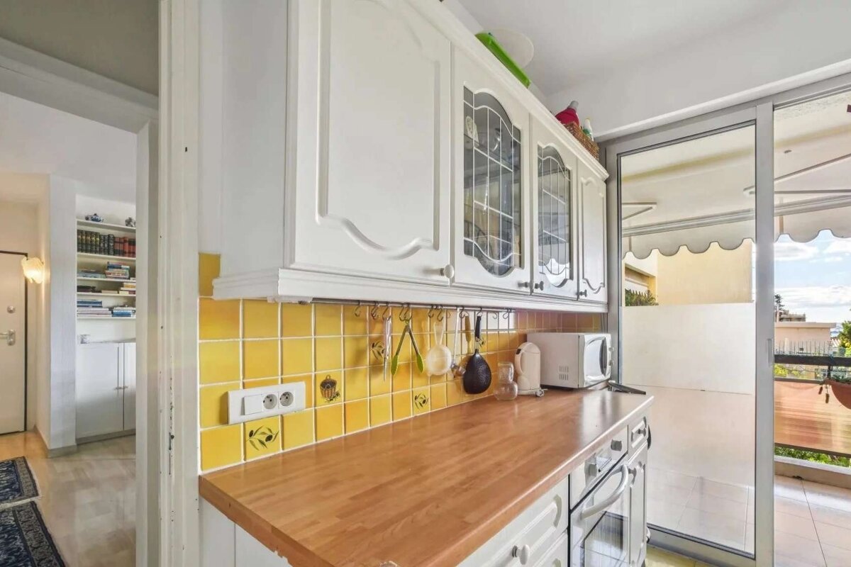 A kitchen with white cabinets, a yellow tiled backsplash, and a wooden counter. It opens to a balcony on the right, with a hallway visible left.