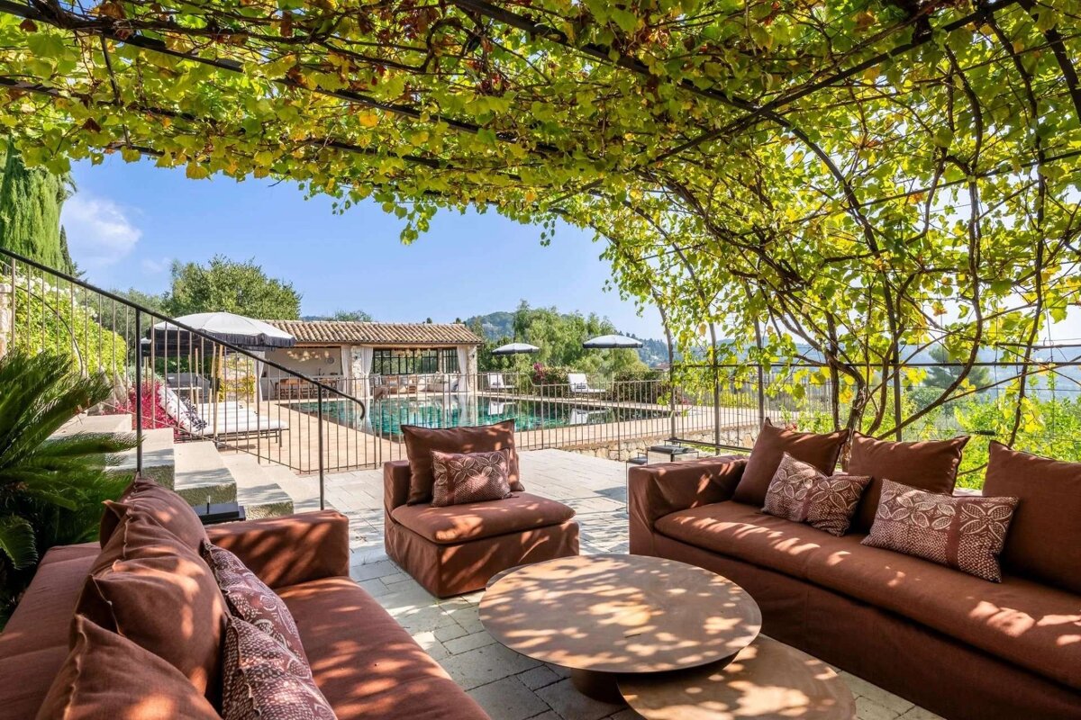 Sunny outdoor lounge with brown furniture shaded by a green vine pergola, revealing a pool and distant hills.