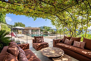 Sunny outdoor lounge with brown furniture shaded by a green vine pergola, revealing a pool and distant hills.