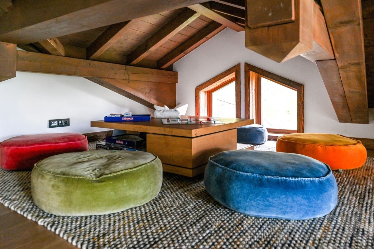 A cozy attic game nook with exposed wooden beams, a low table with board games, and colorful floor poufs on a textured rug.