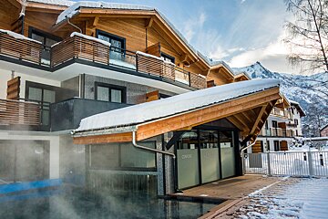 A snowy wooden alpine hotel with balconies and a steaming outdoor pool, framed by majestic snow-capped mountains under a bright sky.