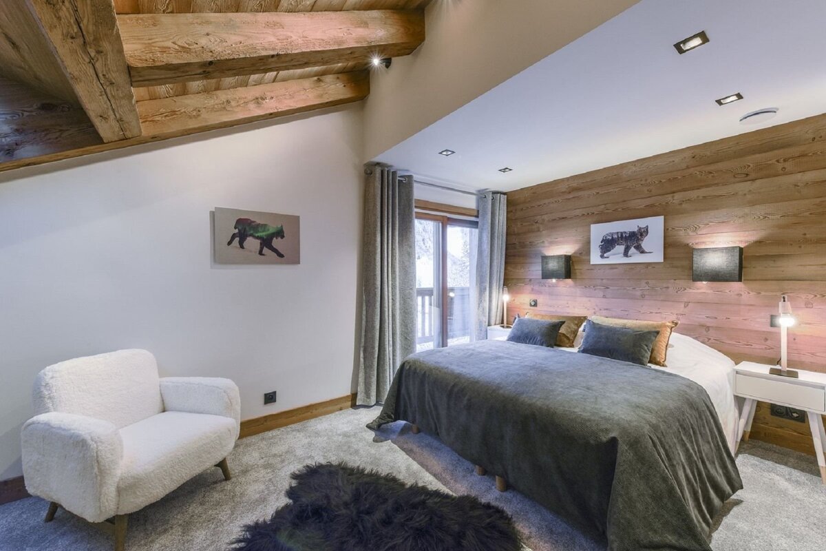 Charming chalet-style bedroom featuring wooden beams, a paneled accent wall, a large bed, plush armchair, and balcony views.