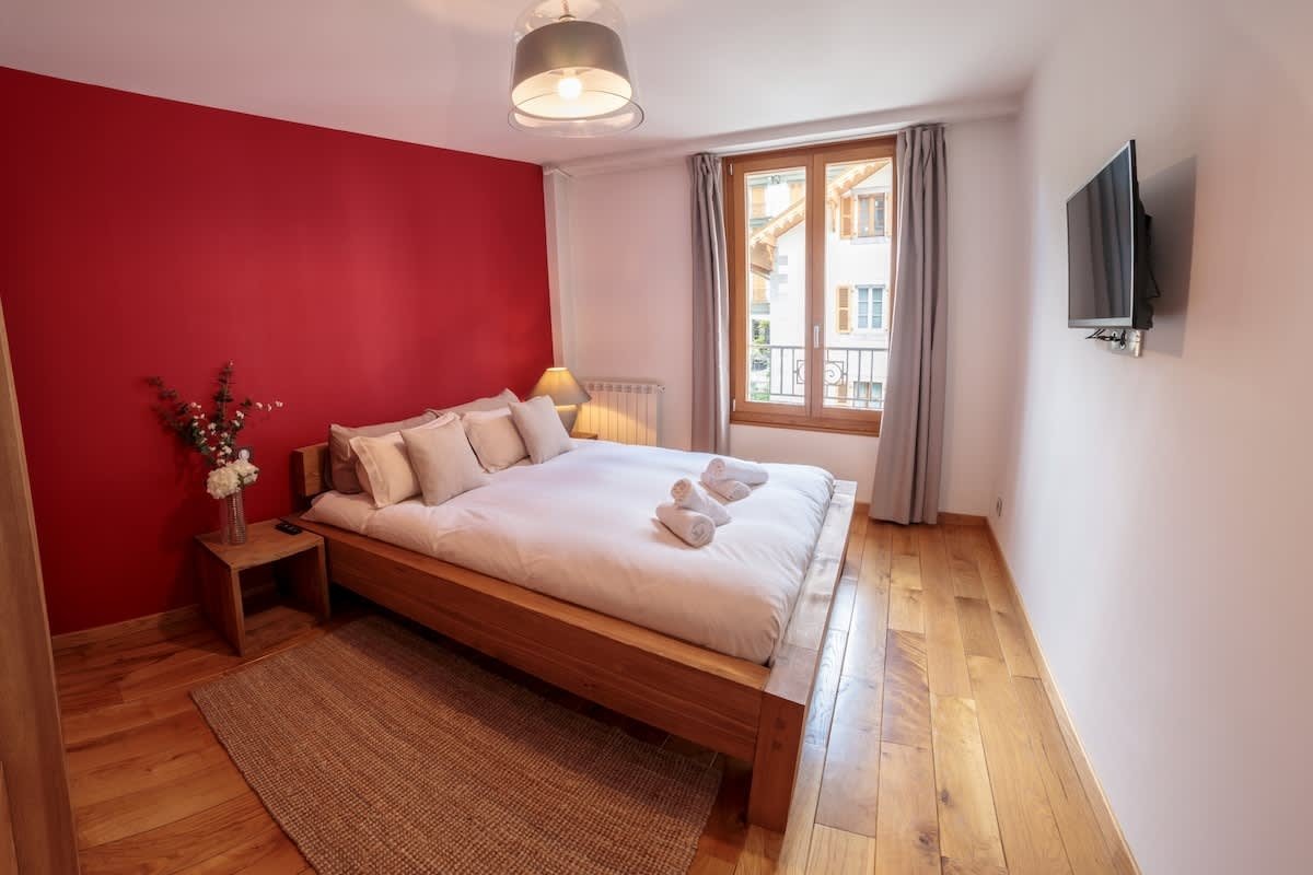 A cozy bedroom with a wooden bed, a vibrant red accent wall, hardwood floors, and a window showing an outdoor view.