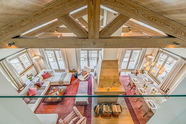 Grand chalet living space seen from above, featuring striking exposed wooden beams, a comfy white sofa, dining area, fireplace, and plush red rugs.