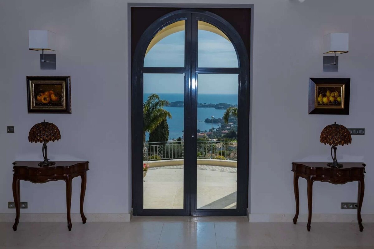 An elegant room with an arched doorway frames a stunning sea and coastal town view. Ornate tables with lamps, paintings, and wall sconces decorate the space.