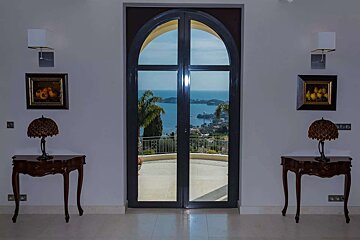 An elegant room with an arched doorway frames a stunning sea and coastal town view. Ornate tables with lamps, paintings, and wall sconces decorate the space.