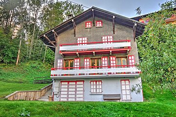 A multi-story chalet with gray walls, striking red trim, and white balconies/shutters, nestled on a lush green hillside amidst trees.