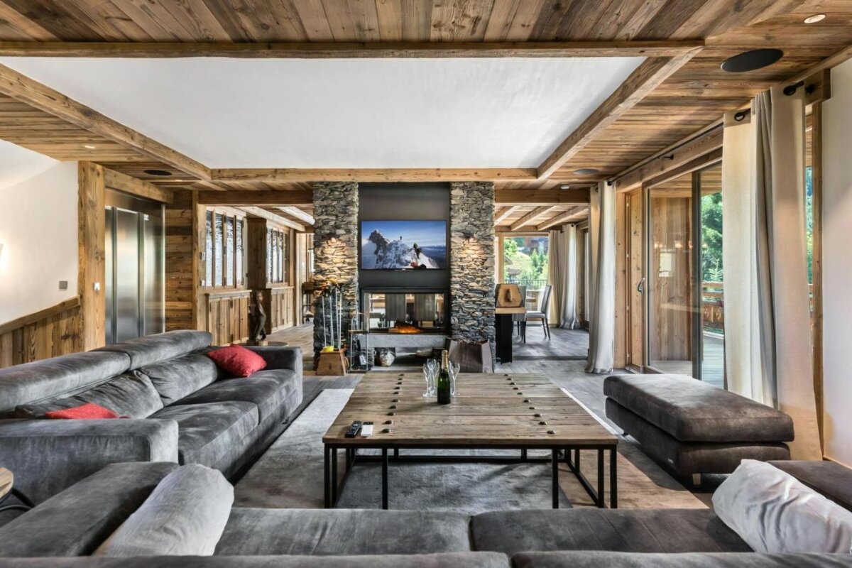 A luxurious rustic-modern living room with exposed wood beams, a stone fireplace, large gray sectional sofas, and a wooden coffee table.