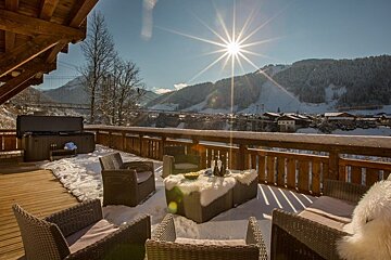 A sunny, snowy chalet balcony with cozy wicker seating, a hot tub, and beautiful views of ski slopes and mountains.