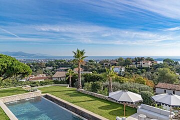 Stunning infinity pool and green lawn with palm trees, offering panoramic views of a coastal town, distant sea, and mountains under a bright blue sky.