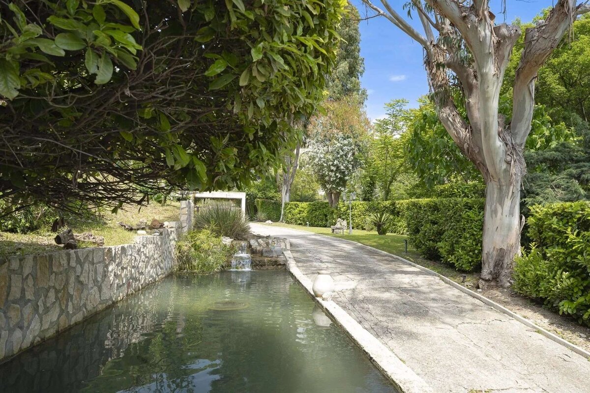 A lush green garden with a paved path alongside a narrow canal featuring a small waterfall, surrounded by trees and hedges under a bright sky.