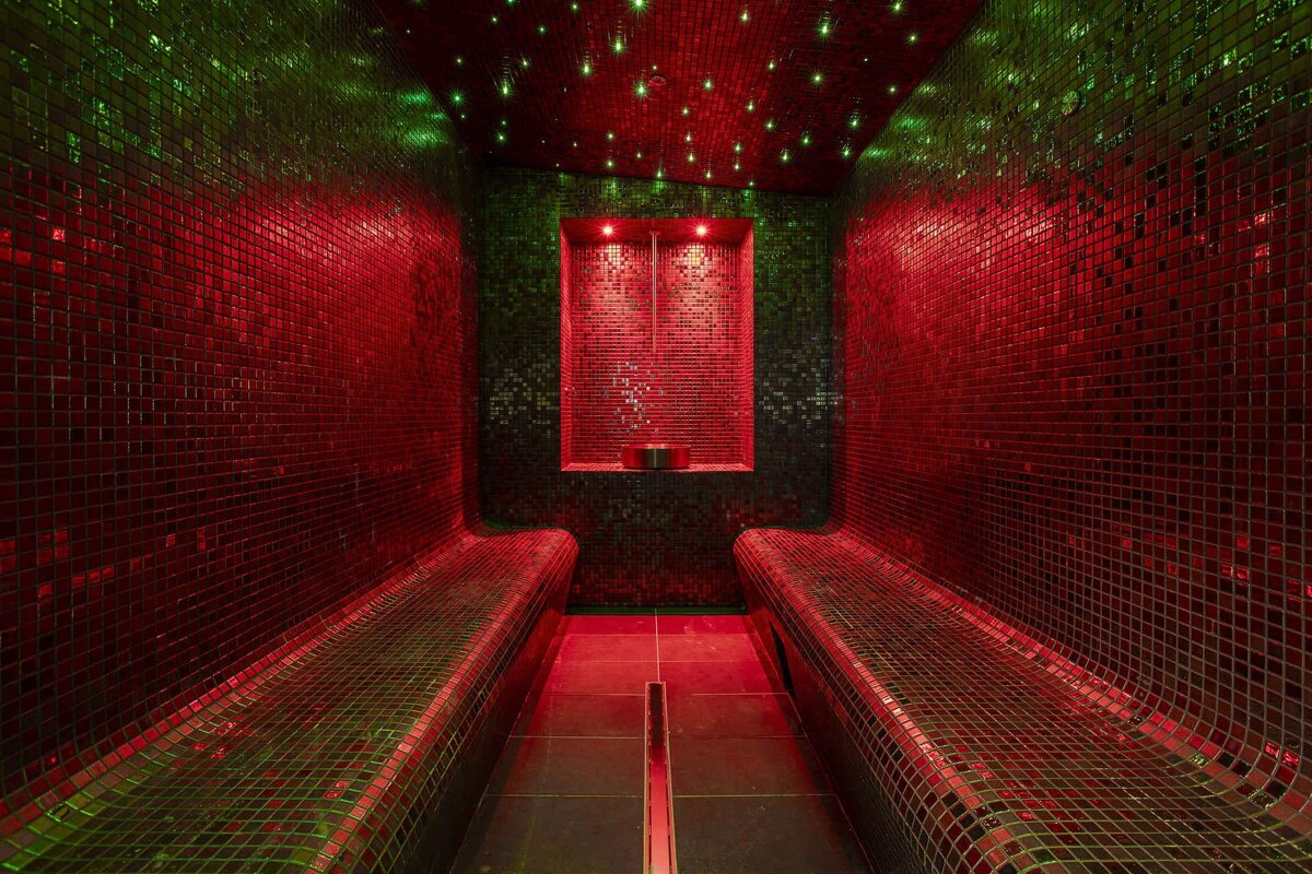 A mosaic-tiled steam room illuminated by vibrant red and green lights. It has two curved benches and twinkling ceiling lights.