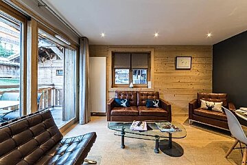Cozy chalet living room with wood-paneled walls, brown leather couches, a glass coffee table, and large windows.