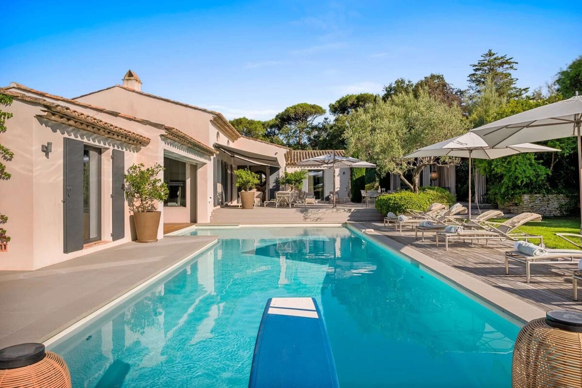 A luxurious villa with a sparkling blue swimming pool, sun loungers, and lush green landscaping under a clear sky.