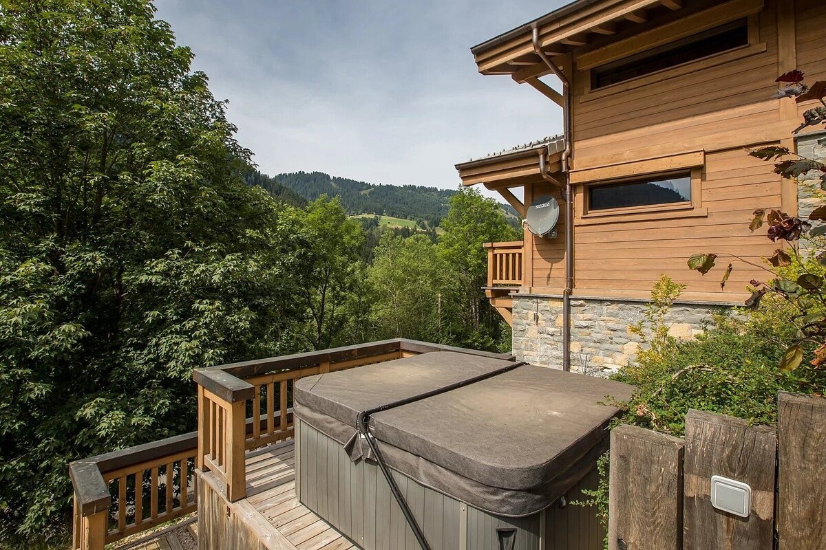 A hot tub on a wooden deck next to a rustic chalet, overlooking a lush green mountain forest under a cloudy sky.