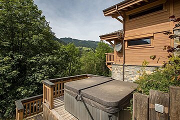 A hot tub on a wooden deck outside a rustic chalet, overlooking a lush green valley and forested mountains under a cloudy sky.