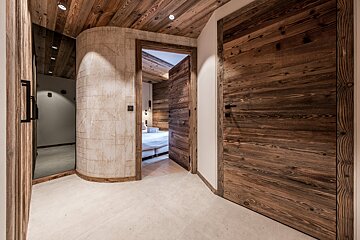 Rustic-modern hallway with wooden doors and ceiling, a curved wall, and an open doorway leading to a cozy bedroom.