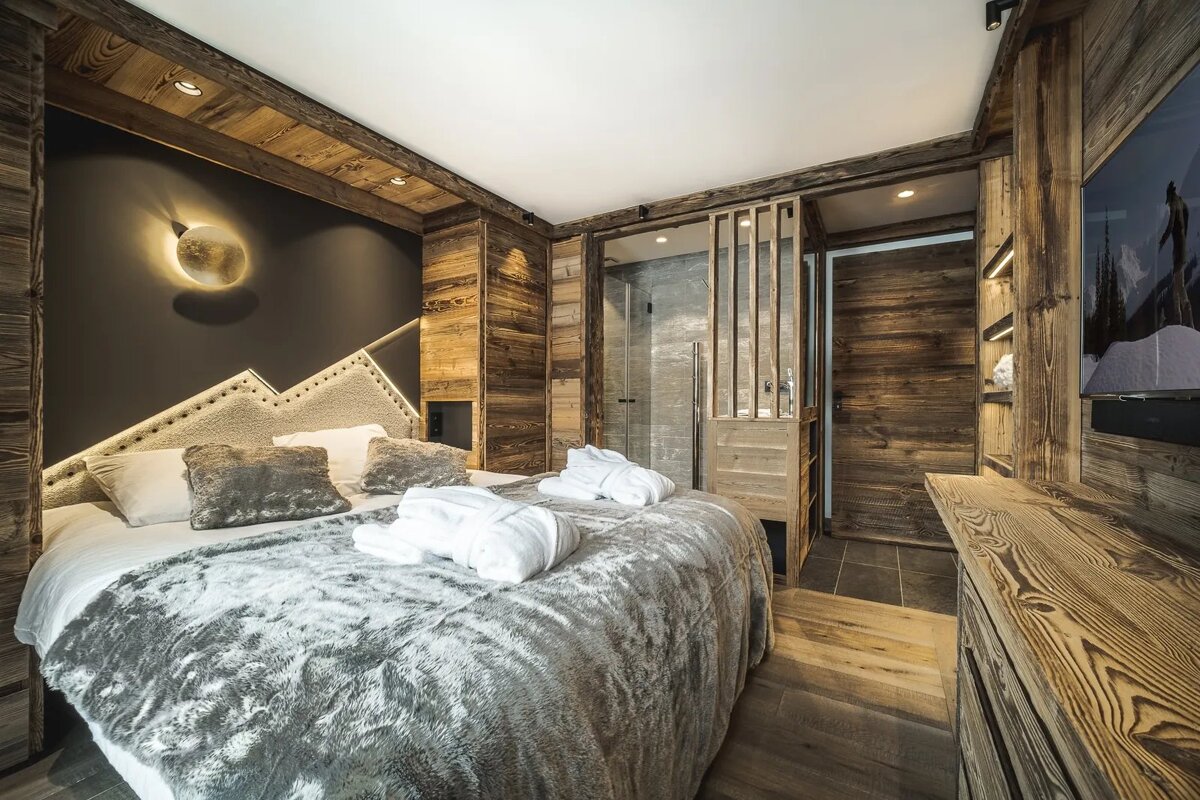 Rustic chalet bedroom with wooden walls, a mountain-shaped headboard, a plush grey fur blanket, and an en-suite bathroom.