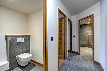 A modern hallway with white walls, wooden door frames, and gray tiled floors. A wall-mounted toilet is on the left, with another bathroom visible ahead.
