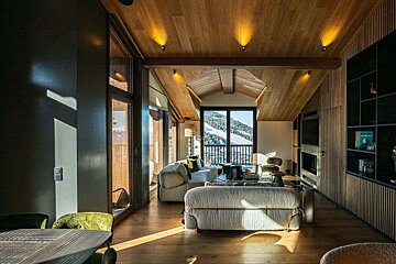 Cozy, modern chalet living room with warm wooden ceilings, plush sofas, and a large window looking out onto snowy mountain slopes.
